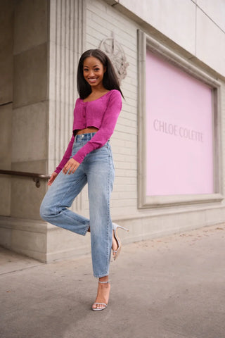 The model is wearing a fuchsia lite knit sweater with blue jeans walking on a side walk with a Chloe Colete pink window sign
